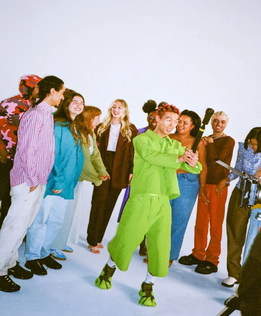 Un groupe de personnes se tenant ensemble dans un studio, avec une personne en tenue vert vif tenant un appareil photo ou un objet similaire, et d'autres personnes souriant et interagissant autour d'eux.