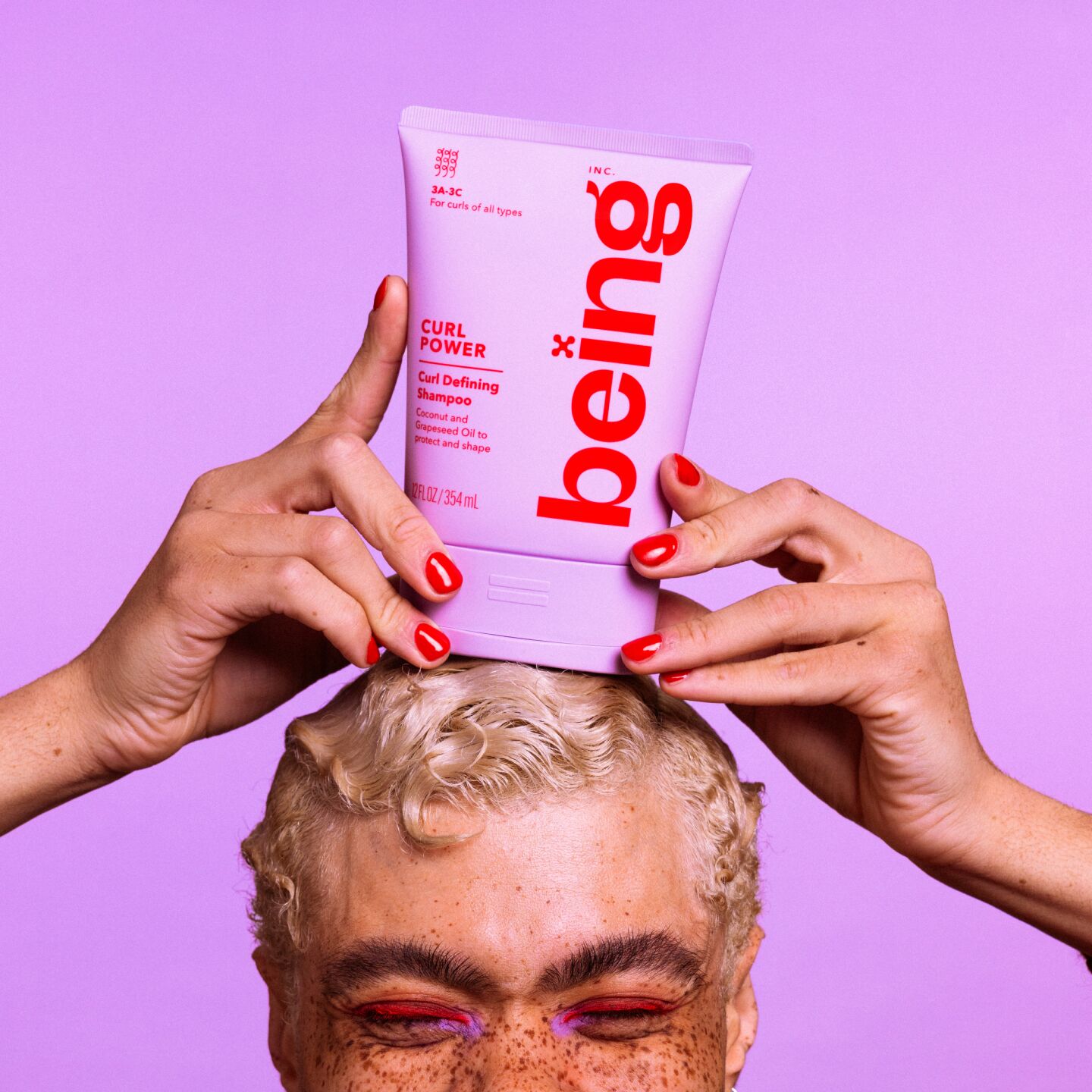 Una persona con pelo rubio corto y pecas sostiene sobre su cabeza un frasco de color lavanda de "ser" Curl Power Curl Defining Shampoo, sobre un fondo de color lavanda.