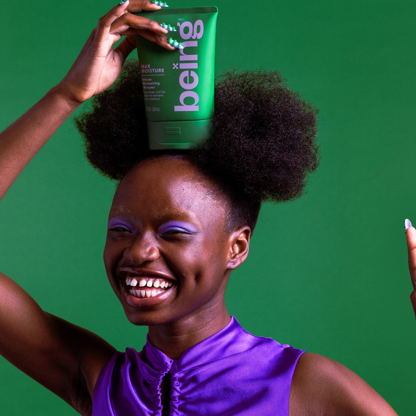 Una persona sonriente, de piel oscura y con dos pelucas afro, sostiene en la cabeza un frasco verde de champú hidratante intenso "being" Max Moisture, sobre un fondo verde. Lleva un top morado y sombra de ojos morada, con las uñas pintadas de verde y blanco.