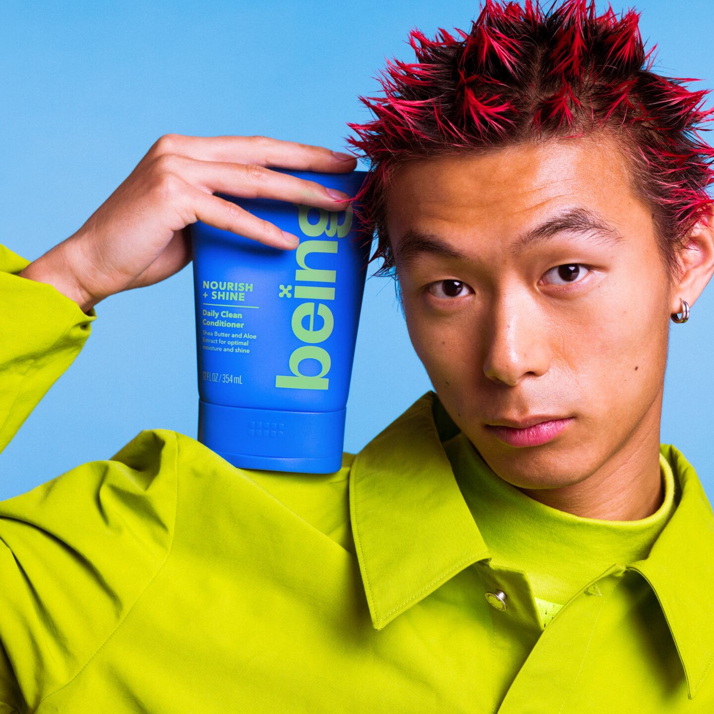 Una persona pelirroja sostiene contra su cabeza un frasco azul de acondicionador Nourish + Shine Daily Clean. Lleva un conjunto verde sobre un fondo azul.