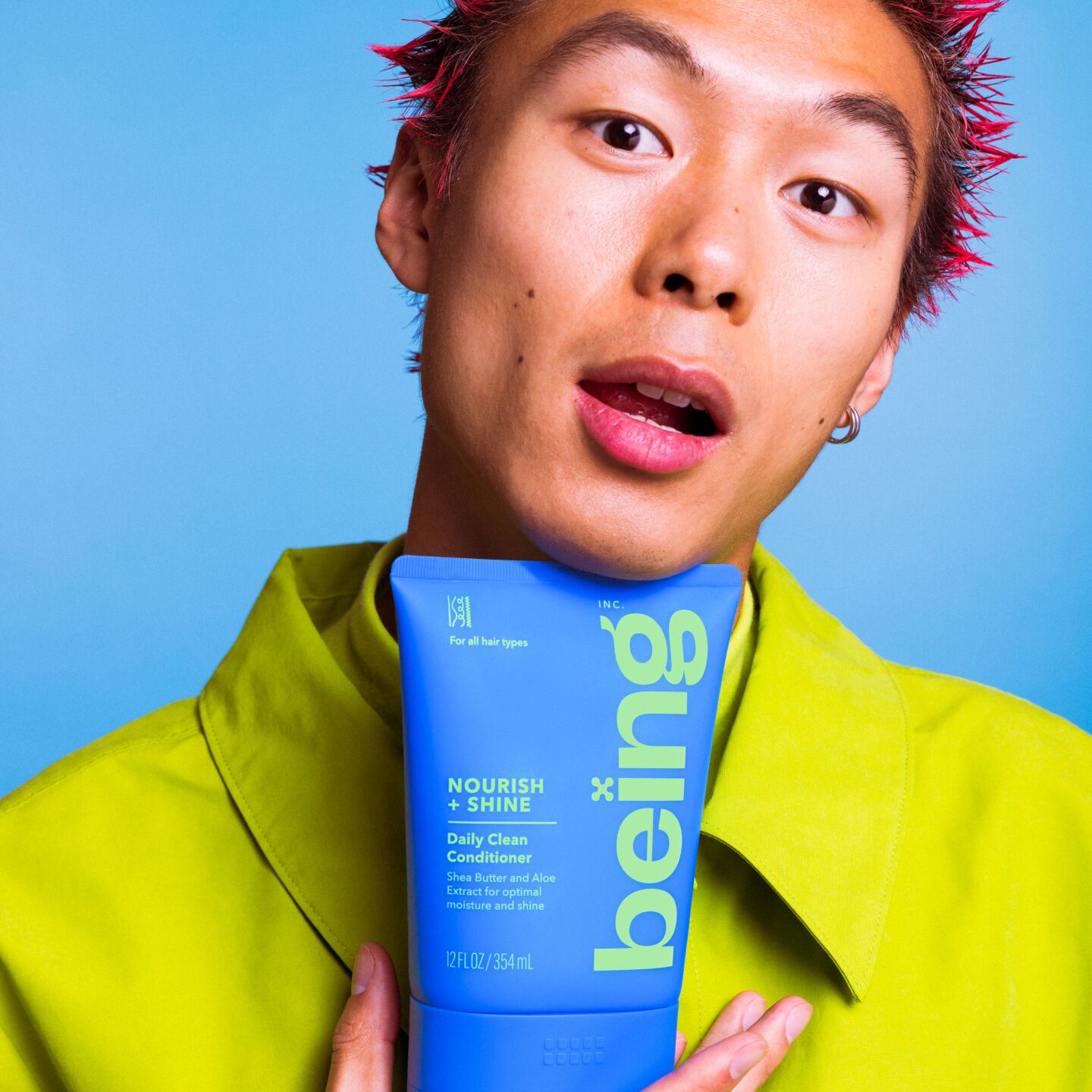 Una persona pelirroja con el pelo de punta sostiene contra el pecho un frasco azul de "ser" Acondicionador Nourish + Shine Daily Clean. Lleva un conjunto verde sobre un fondo azul.