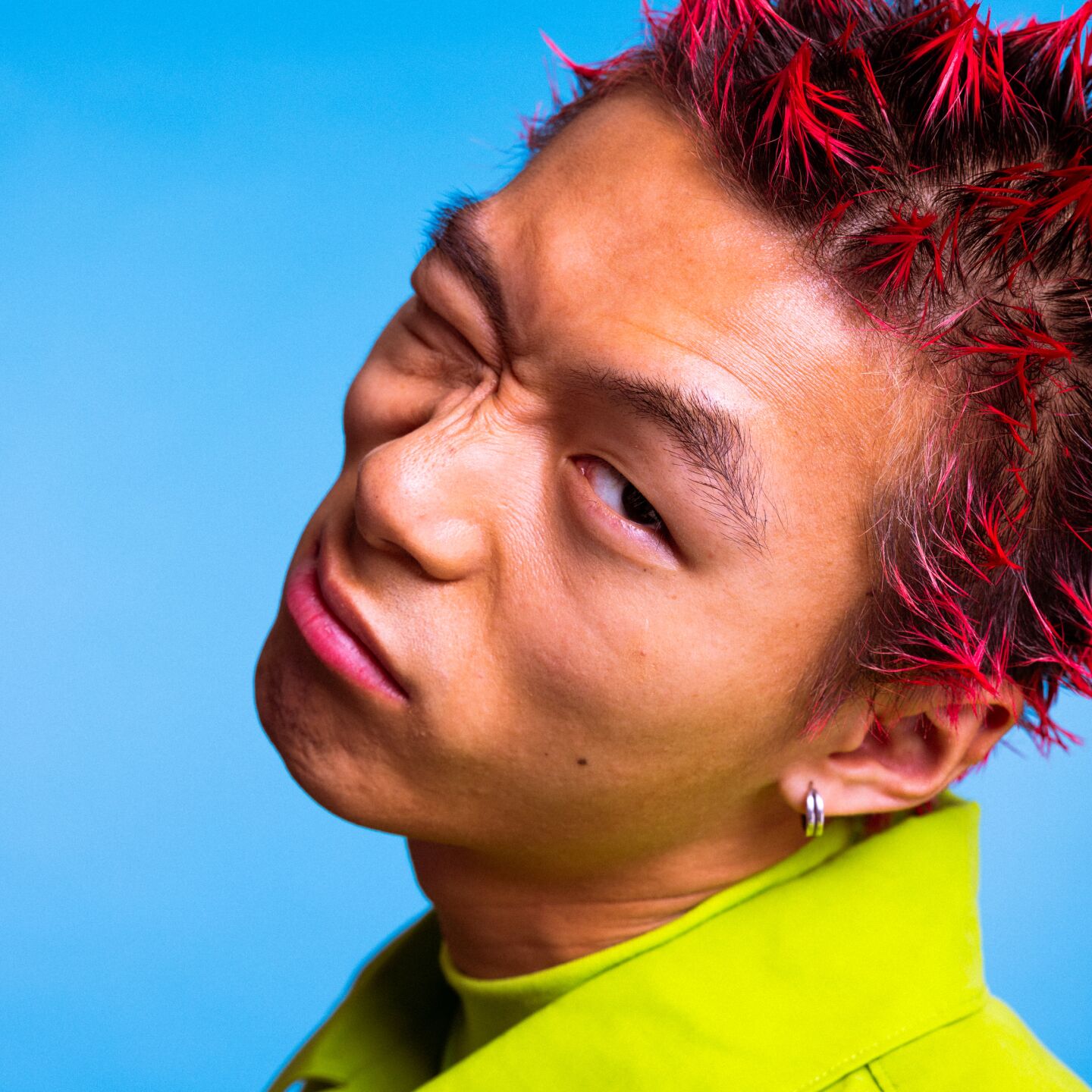 Una persona con el pelo pelirrojo de punta hace una expresión facial crispada, con un traje verde, sobre un fondo azul.